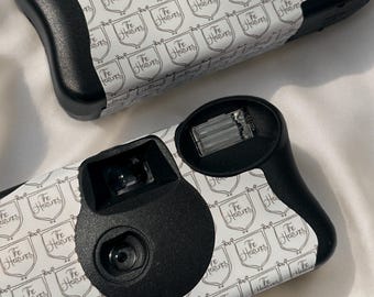 Puede incluir: Dos cámaras desechables en blanco y negro. Las cámaras tienen un patrón repetido de escudos con el texto "The Heavens". Tienen empuñaduras y lentes negras.