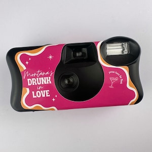 May include: A black disposable camera with a pink and orange design. The camera has a sticker that says "Montana's Drunk in Love" with stars and a martini glass icon. The text "press here for flash" is also on the sticker.