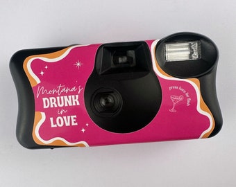 Puede incluir: Una cámara desechable negra con un diseño rosa y naranja. La cámara tiene una pegatina que dice "Montana's Drunk in Love" con estrellas y un icono de copa de martini. El texto "press here for flash" también está en la pegatina.
