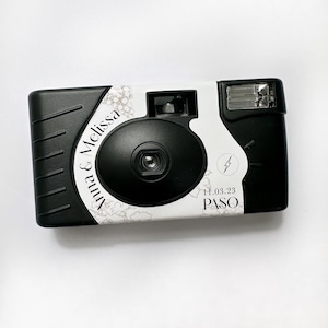 Peut inclure: Un appareil photo jetable noir et blanc avec un motif floral et le texte "Anna & Melissa 11.03.23 PASO" imprimé sur le devant.