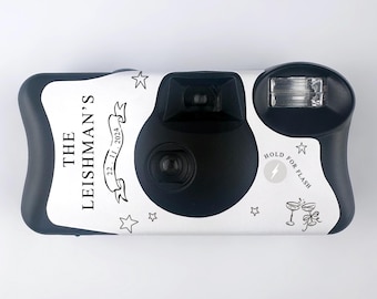 Puede incluir: Cámara desechable blanca con detalles negros y una etiqueta personalizada que dice "The Leishman's 22.11.2024". La cámara tiene un flash y un botón de retención del flash.