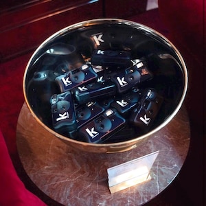 May include: A black bowl filled with several small, black cameras. Each camera has a white "k" logo. The bowl is gold-rimmed and sits on a marble-patterned surface. A small, angled sign is in front of the bowl.