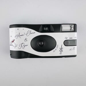 May include: A white and black disposable camera with the text "Anna Claire & Ryan" and the date "28.08.2021" printed on it. The camera has a flash and a small viewfinder.