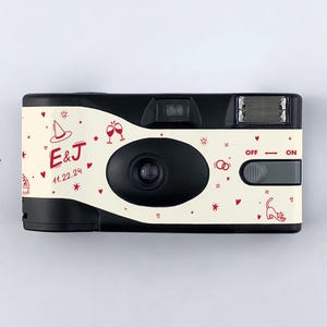 May include: A black and white disposable camera with a white background. The camera has a red and white design with hearts, stars, and other whimsical illustrations. The text "E&J 11.22.24" is printed on the camera.
