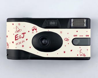 May include: A black and white disposable camera with a white background. The camera has a red and white design with hearts, stars, and other whimsical illustrations. The text "E&J 11.22.24" is printed on the camera.