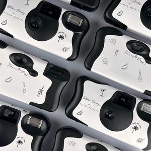 May include: A set of eight disposable cameras with black and white cow print designs. Each camera features a different design with text that reads "Turn on the flash!" and "Blake Healey x Status Anxiety."