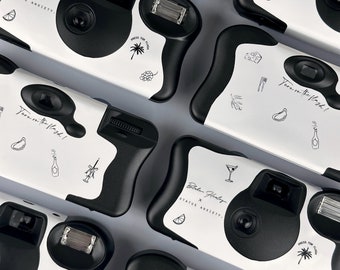 Puede incluir: Un conjunto de ocho cámaras desechables con diseños de vaca en blanco y negro. Cada cámara presenta un diseño diferente con texto que dice "¡Enciende el flash!" y "Blake Healey x Status Anxiety."
