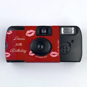 May include: Red disposable camera with the text "Lara's 30th Birthday" and pink lips printed on the front. The camera has a black flash and a date window that says "December 2024".