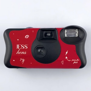 May include: A red disposable camera with black accents. The camera has the text "JESS hens" and "press for flash" printed on it, along with small decorative icons. The camera is designed for taking photos.