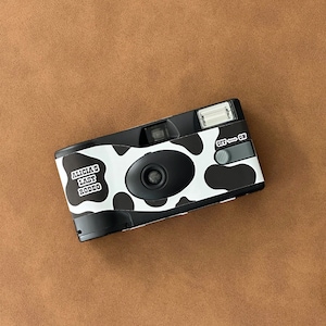 May include: A black and white disposable camera with a cow print design. The camera has the text "Alicia's Last Rodeo" printed on it.