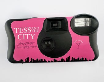Puede incluir: Cámara desechable rosa con lente negra y flash. La cámara tiene un diseño con el texto "Tess and the City" y una silueta de ciudad. El texto "And just like that Tess is getting married!" también está incluido en la cámara.