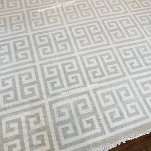May include: A close-up of a light gray and white patterned rug. The rug features a repeating geometric design with a Greek key pattern. The rug has a fringed edge and the text "Noor Handloom Carpets" is visible.