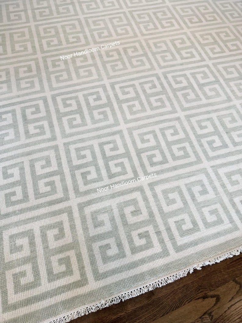 May include: A close-up of a light gray and white patterned rug. The rug features a repeating geometric design with a Greek key pattern. The rug has a fringed edge and the text "Noor Handloom Carpets" is visible.