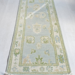 May include: A rectangular, light blue rug with a floral pattern and a cream-colored border. The rug has a green and cream-colored floral design in the center and along the border. The rug has a fringe on the short ends. The text "NOORHANDLOOMCARPETS" is visible on the rug.