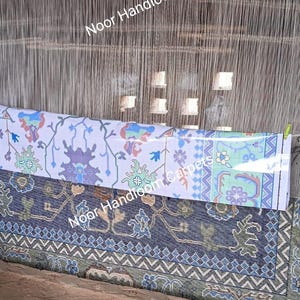 May include: A handloom weaving setup with a partially completed carpet. The carpet features intricate floral and geometric patterns in shades of blue, green, and purple on a white and navy background. The text "Noor Handloom Carpets" is visible.