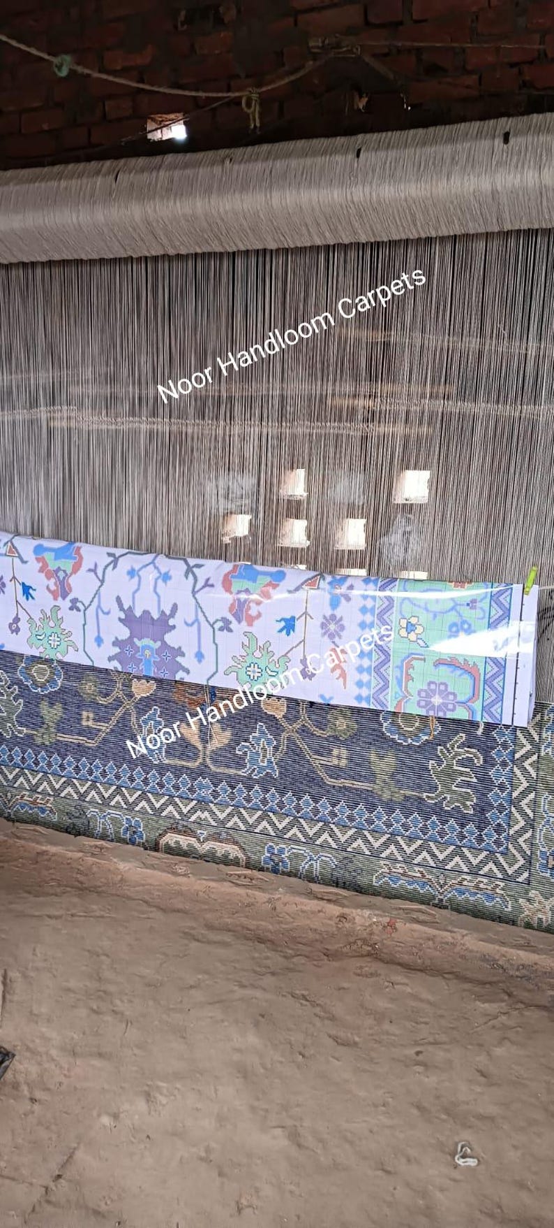 May include: A handloom weaving setup with a partially completed carpet. The carpet features intricate floral and geometric patterns in shades of blue, green, and purple on a white and navy background. The text "Noor Handloom Carpets" is visible.