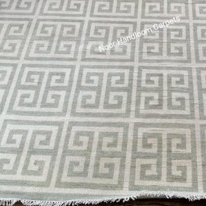 May include: A light gray and white geometric patterned rug with a Greek key design. The rug has a textured appearance and a fringed edge. The text "Noor Handloom Carpets" is visible on the rug.