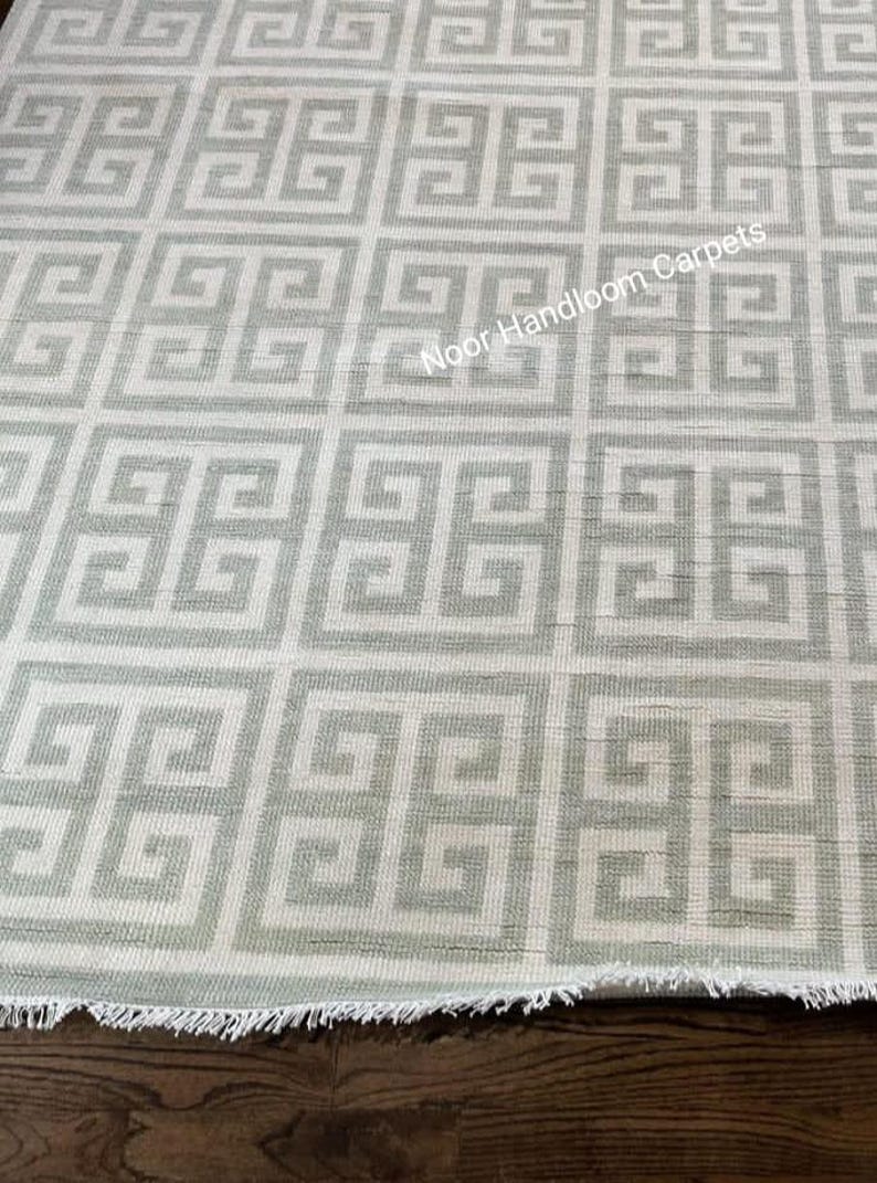 May include: A light gray and white geometric patterned rug with a Greek key design. The rug has a textured appearance and a fringed edge. The text "Noor Handloom Carpets" is visible on the rug.