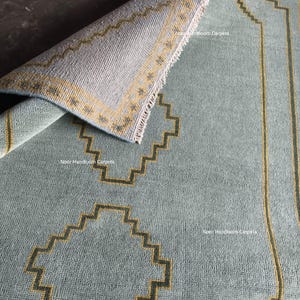 May include: A light blue area rug with a folded corner revealing the underside. The rug features a dark yellow geometric stepped pattern and the text Noor Handloom Carpets printed in white. The edges have a short white fringe detail.