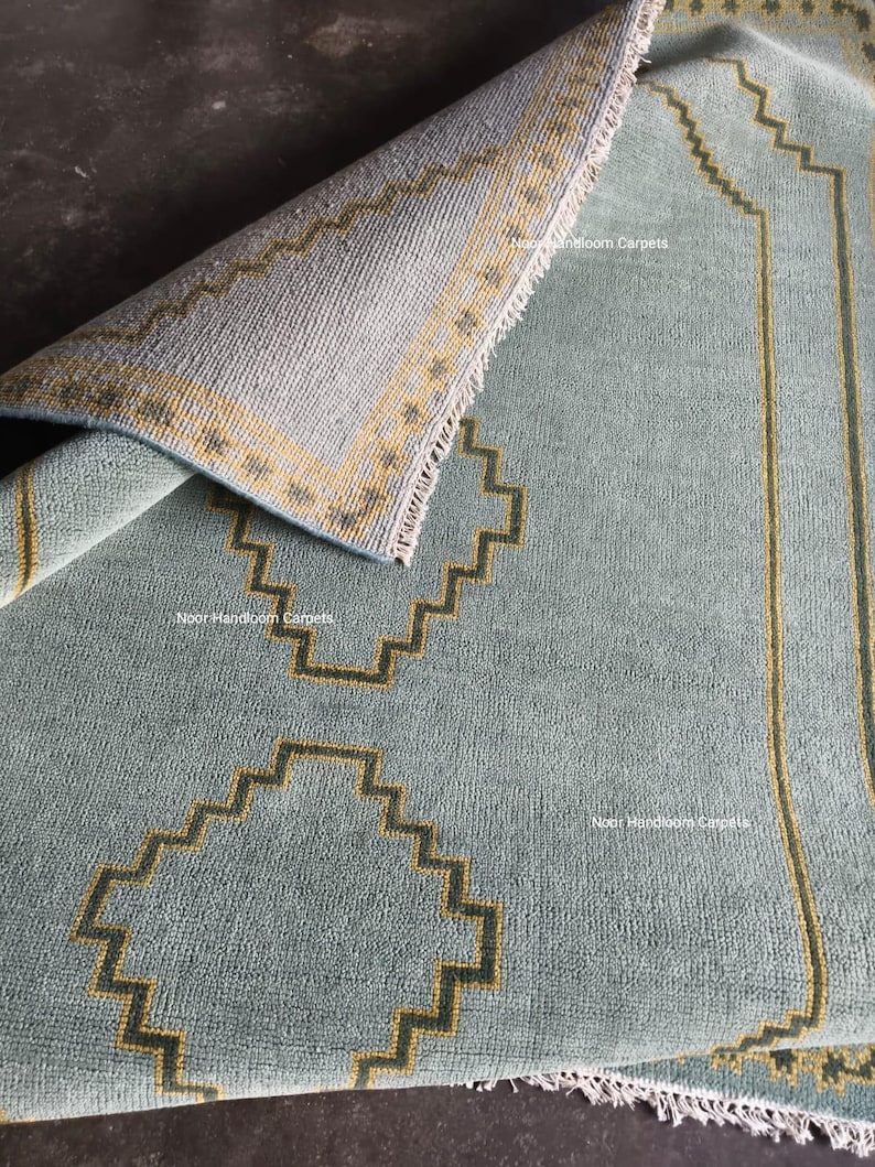 May include: A light blue area rug with a folded corner revealing the underside. The rug features a dark yellow geometric stepped pattern and the text Noor Handloom Carpets printed in white. The edges have a short white fringe detail.