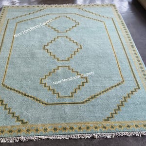 May include: A rectangular area rug in light blue with a mustard yellow border featuring a repeating star pattern. The center displays a large octagonal outline and three stacked diamond shapes in mustard yellow, with the text Noor Handloom Carpets.