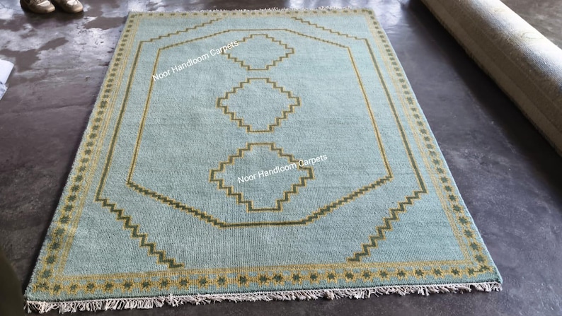 May include: A rectangular area rug in light blue with a mustard yellow border featuring a repeating star pattern. The center displays a large octagonal outline and three stacked diamond shapes in mustard yellow, with the text Noor Handloom Carpets.