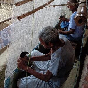 May include: Two men weaving a rug on a traditional loom. The men are seated, working with white threads and wooden tools. The loom has multiple wooden rollers and a woven structure. The scene is lit by natural light.