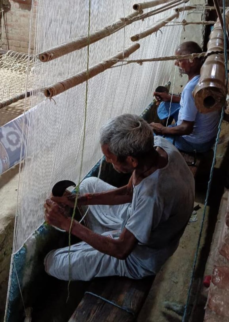 May include: Two men weaving a rug on a traditional loom. The men are seated, working with white threads and wooden tools. The loom has multiple wooden rollers and a woven structure. The scene is lit by natural light.