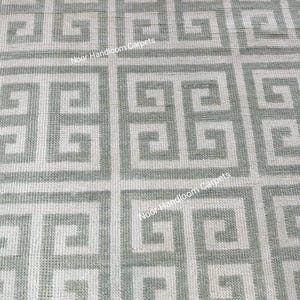 May include: A close-up view of a gray and white patterned rug. The rug features a repeating geometric design with a Greek key pattern. The text "Noor Handloom Carpets" is visible on the rug.