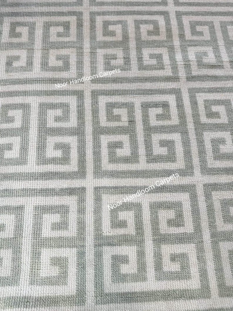 May include: A close-up view of a gray and white patterned rug. The rug features a repeating geometric design with a Greek key pattern. The text "Noor Handloom Carpets" is visible on the rug.