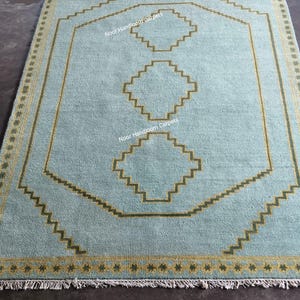 May include: A light blue rectangular area rug featuring a central vertical row of three stepped geometric shapes in olive green. The rug has a matching olive green zigzag border and a fringe on the ends. The text Noor Handloom Carpets is visible.