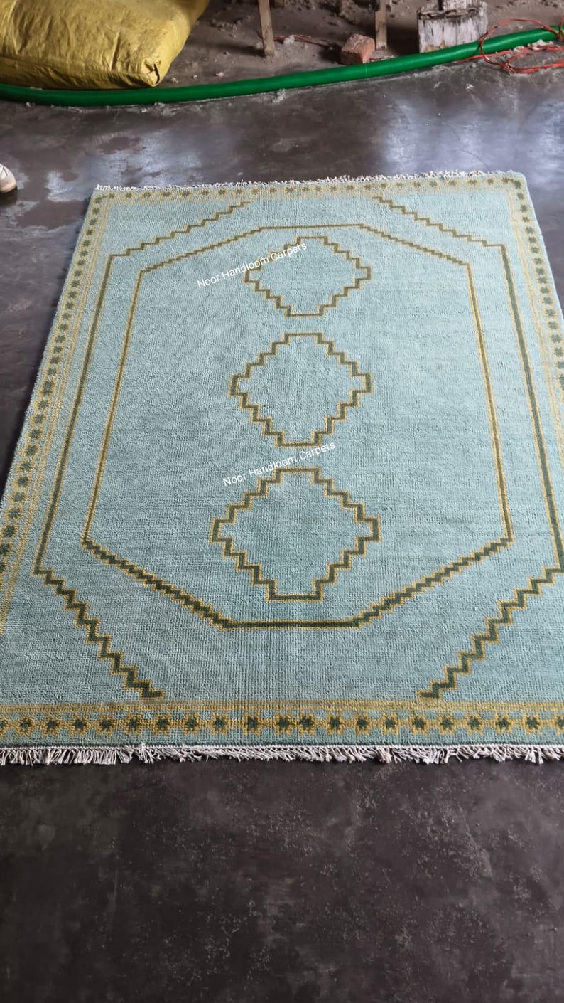 May include: A light blue rectangular area rug featuring a central vertical row of three stepped geometric shapes in olive green. The rug has a matching olive green zigzag border and a fringe on the ends. The text Noor Handloom Carpets is visible.