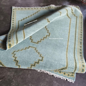 May include: A light blue hand-woven rug featuring a geometric stepped border and central motif in yellow and dark green. The rug has a fringed edge and displays the text Noor Handloom Carpets printed twice on the surface of the fabric.