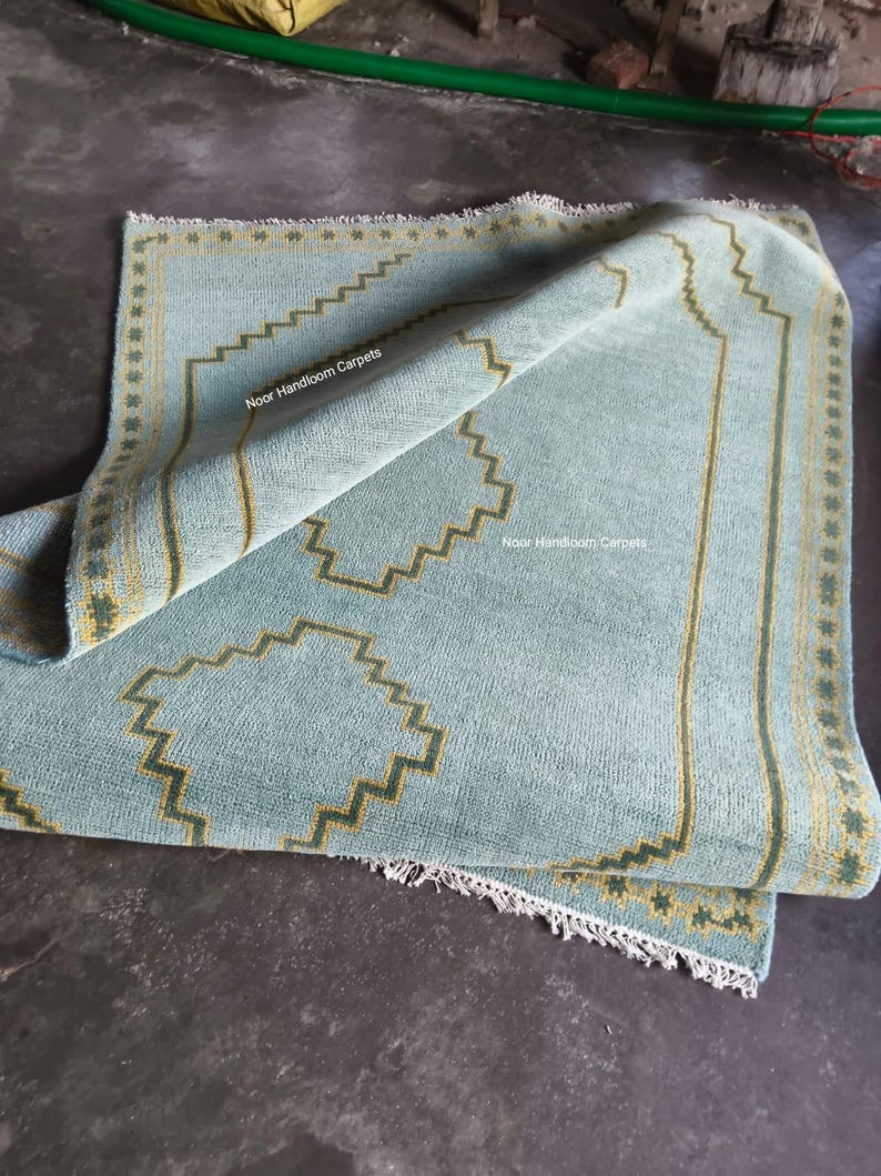 May include: A light blue hand-woven rug featuring a geometric stepped border and central motif in yellow and dark green. The rug has a fringed edge and displays the text Noor Handloom Carpets printed twice on the surface of the fabric.