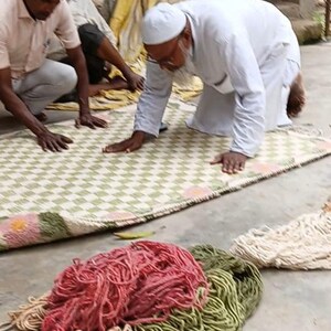 May include: Men working on a woven rug with a green and white checkered pattern. The rug has a pink and green floral border. Bundles of colorful yarn in red, green, and beige are in the foreground.