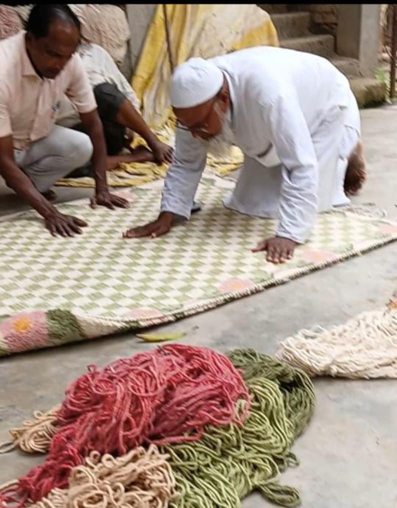 May include: Men working on a woven rug with a green and white checkered pattern. The rug has a pink and green floral border. Bundles of colorful yarn in red, green, and beige are in the foreground.