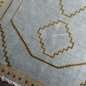 May include: A light blue area rug featuring a dark mustard yellow geometric border and central zigzag motifs. The rug has a textured pile surface, a repeating pattern of dark squares along the edge, and a short white fringe at the bottom end.
