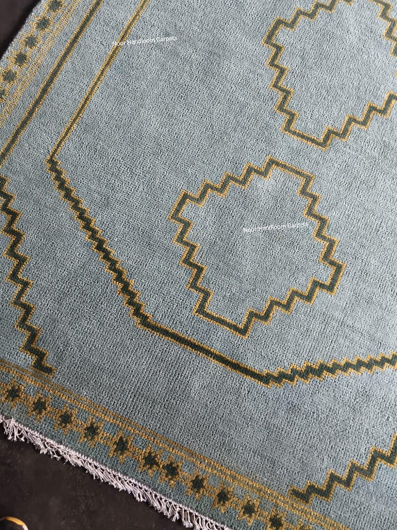 May include: A light blue area rug featuring a dark mustard yellow geometric border and central zigzag motifs. The rug has a textured pile surface, a repeating pattern of dark squares along the edge, and a short white fringe at the bottom end.