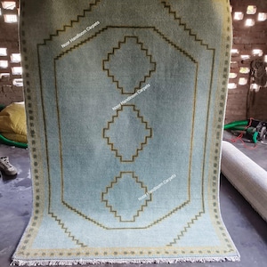 May include: A light blue rectangular rug featuring a dark olive green geometric border and three stacked diamond-shaped motifs in the center. The rug includes a repeating star pattern along the edges and the text Noor Handloom Carpets.