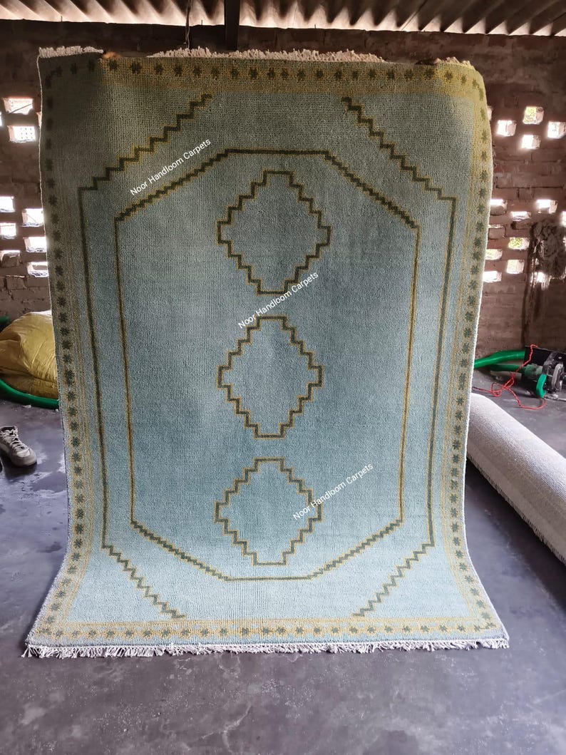 May include: A light blue rectangular rug featuring a dark olive green geometric border and three stacked diamond-shaped motifs in the center. The rug includes a repeating star pattern along the edges and the text Noor Handloom Carpets.