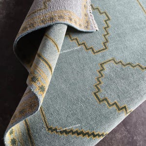 May include: A light blue area rug with a folded corner revealing the underside. The rug features a repeating geometric pattern in yellow and dark green, with a border design. The text Noor Handloom Carpets is visible on the surface.