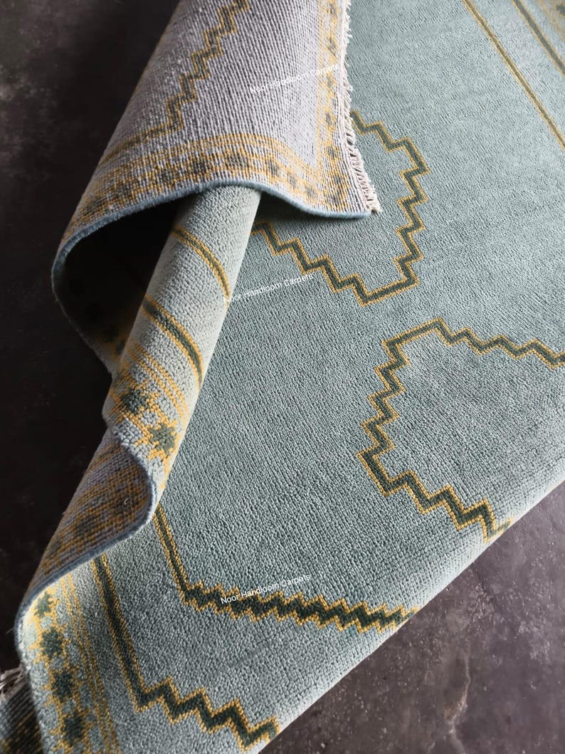 May include: A light blue area rug with a folded corner revealing the underside. The rug features a repeating geometric pattern in yellow and dark green, with a border design. The text Noor Handloom Carpets is visible on the surface.