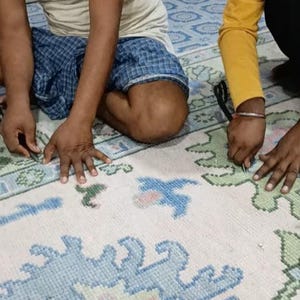 May include: A large, off-white area rug featuring a repeating pattern of stylized floral and geometric motifs in shades of light blue and sage green. The rug has a thick, tufted pile texture and is being worked on by two sets of hands using scissors.