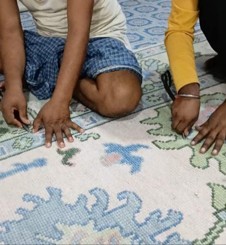 May include: A large, off-white area rug featuring a repeating pattern of stylized floral and geometric motifs in shades of light blue and sage green. The rug has a thick, tufted pile texture and is being worked on by two sets of hands using scissors.
