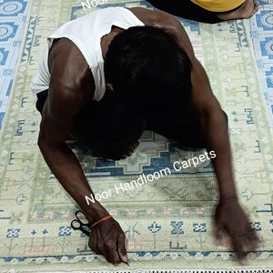 May include: A handmade rug with a light green and blue geometric pattern. The rug has a fringe on the edge. The text "Noor Handloom Carpets" is visible. Two people are working on the rug.