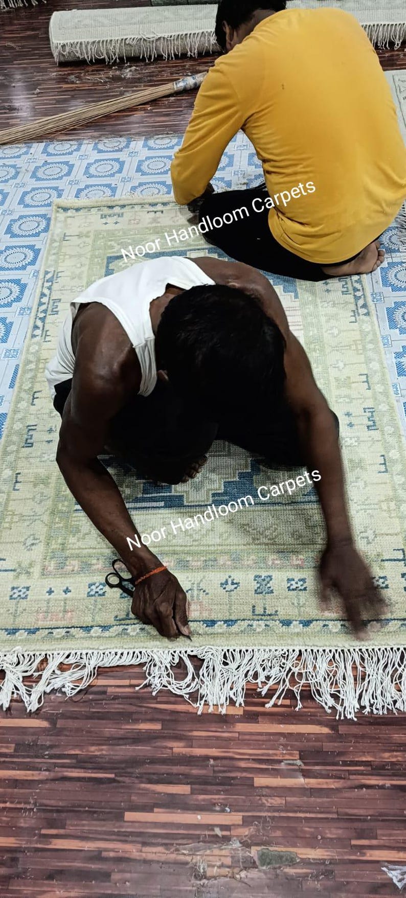 May include: A handmade rug with a light green and blue geometric pattern. The rug has a fringe on the edge. The text "Noor Handloom Carpets" is visible. Two people are working on the rug.