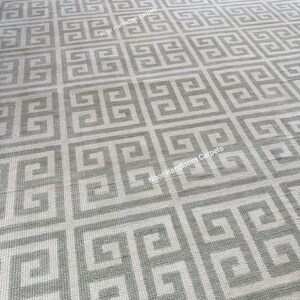 May include: A large, rectangular rug with a repeating geometric pattern in shades of gray and off-white. The design features a Greek key motif, creating a symmetrical and textured surface. The text "Noor Handloom Carpets" is visible on the rug.