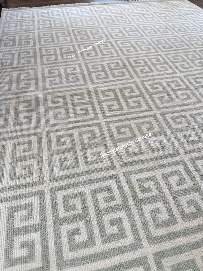 May include: A large, rectangular rug with a repeating geometric pattern in shades of gray and off-white. The design features a Greek key motif, creating a symmetrical and textured surface. The text "Noor Handloom Carpets" is visible on the rug.