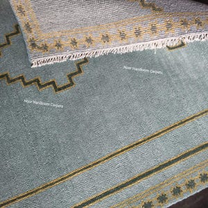 May include: A light blue area rug with a folded corner revealing a textured underside. The rug features dark green and yellow geometric zigzag borders and linear patterns. The text Noor Handloom Carpets is printed on the surface of the rug.