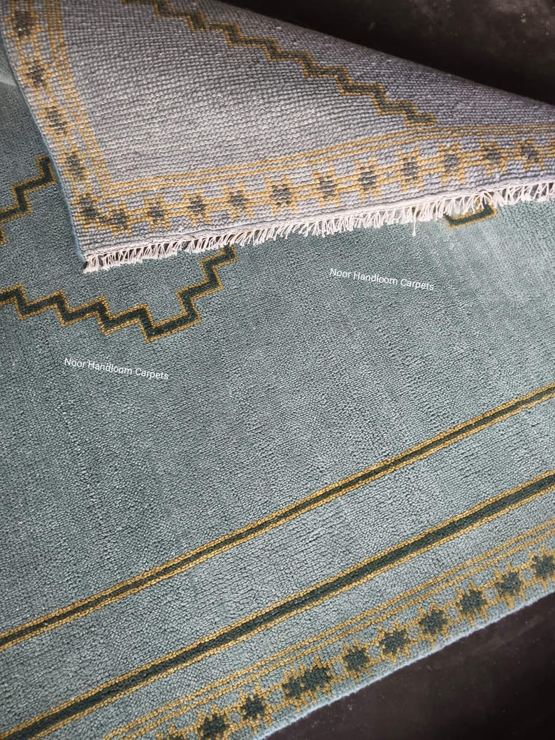 May include: A light blue area rug with a folded corner revealing a textured underside. The rug features dark green and yellow geometric zigzag borders and linear patterns. The text Noor Handloom Carpets is printed on the surface of the rug.